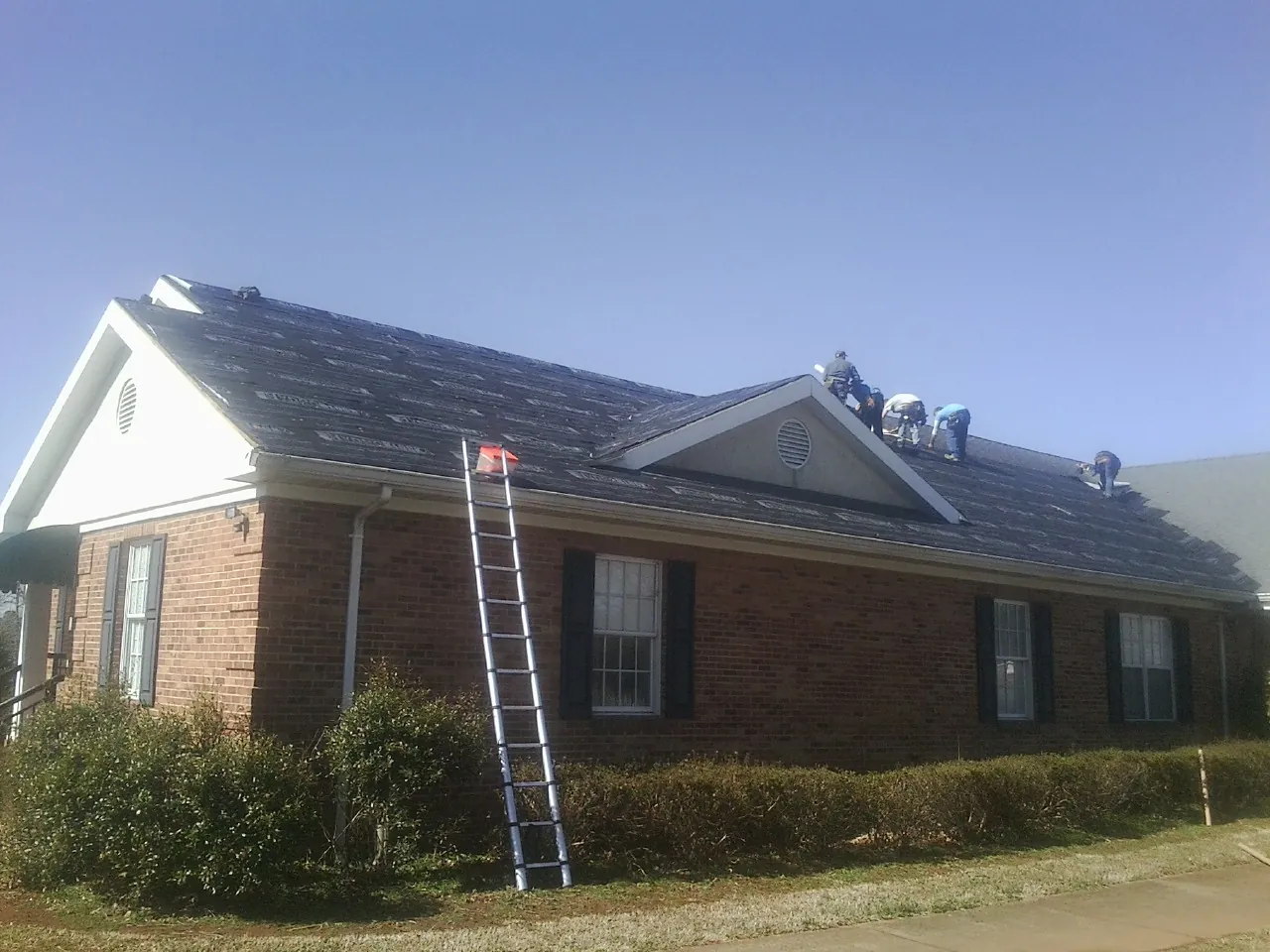 Crew tearing shingles off a brick ranch roof with a ladder set — Metal Roof Repair work by our crew in Crystal Spring