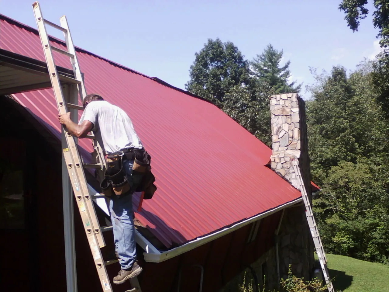 Roofer on a ladder beside a red metal roof and stone chimney — Soffit & Fascia Repair by Roofing Brothers in Crystal Spring